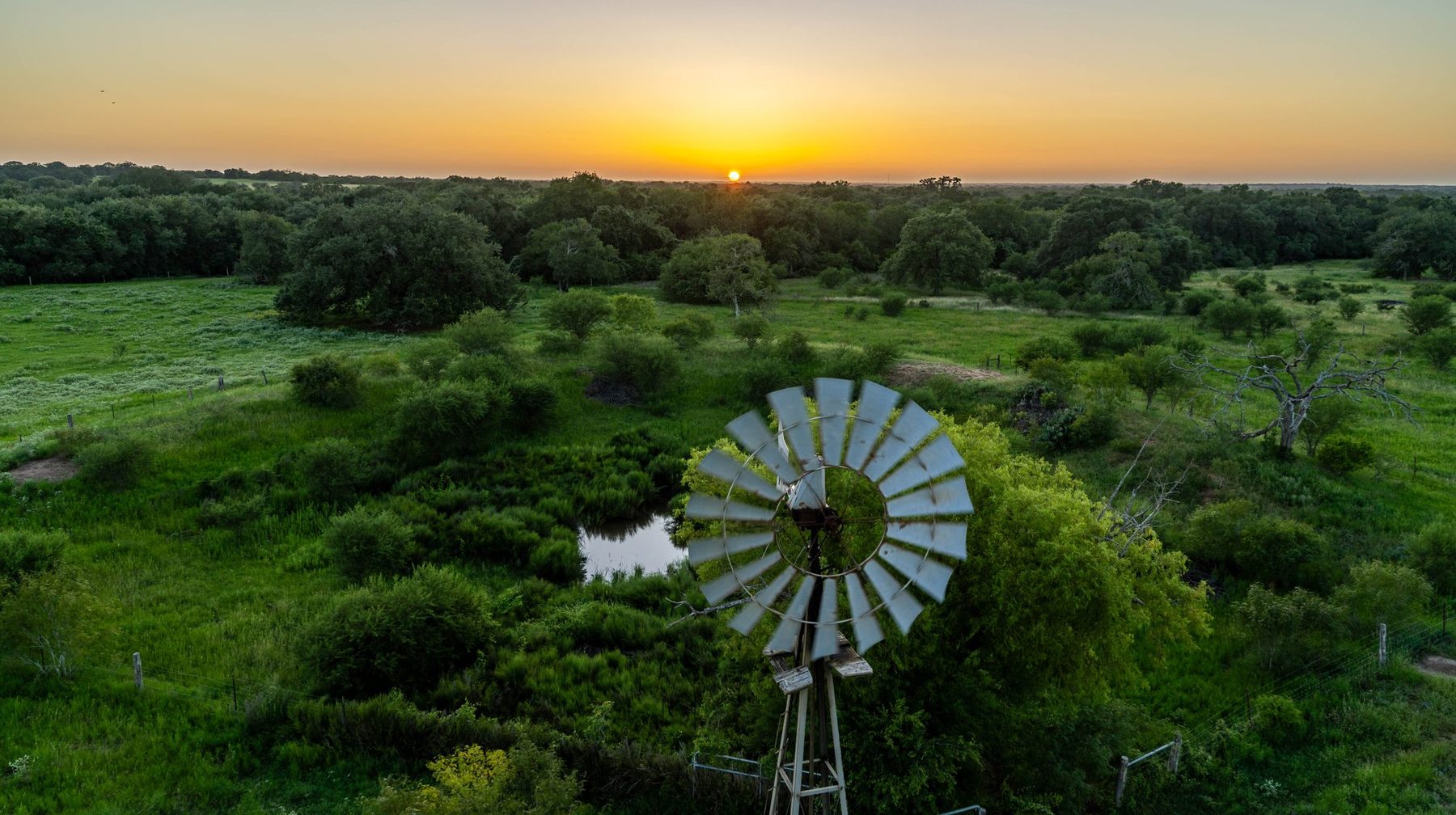 Aerial sunset over a working Texas ranch — windmill, ponds and mature oak pasture, captured by Aerialcam Media